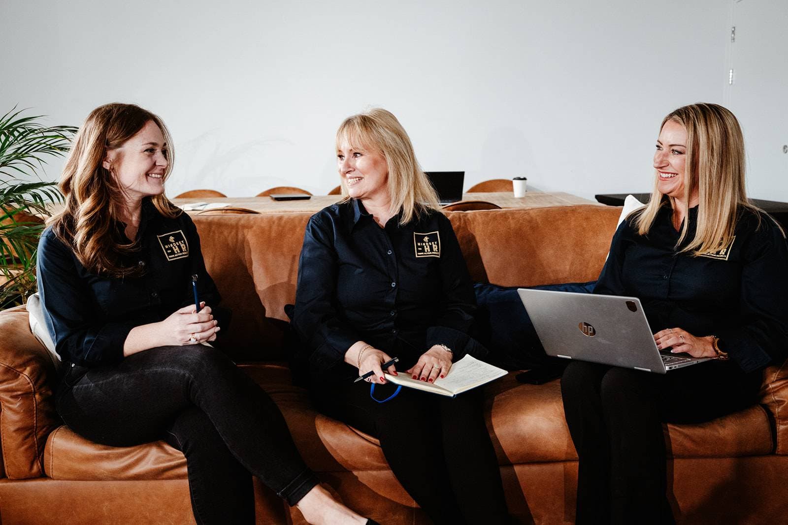 Three Mindset HR consultants in branded shirts talking together on a sofa, one taking notes and one with a laptop