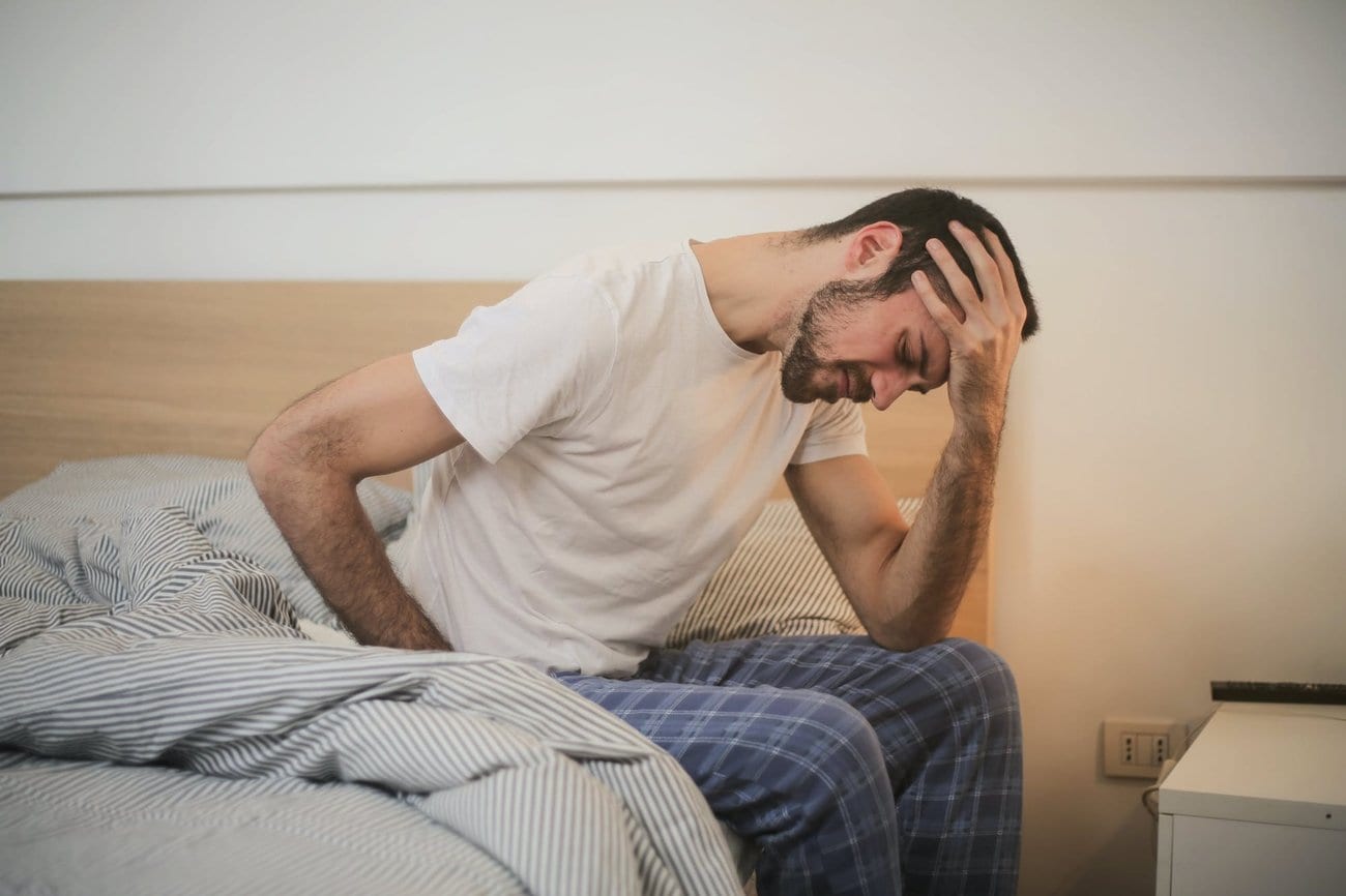 A man sitting on the edge of a bed, holding his head and his lower back