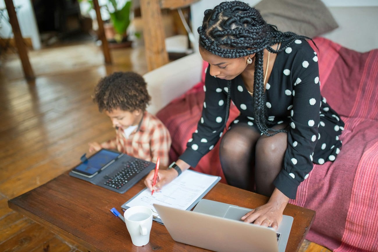 A woman working at a laptop at home while her young son draws beside her